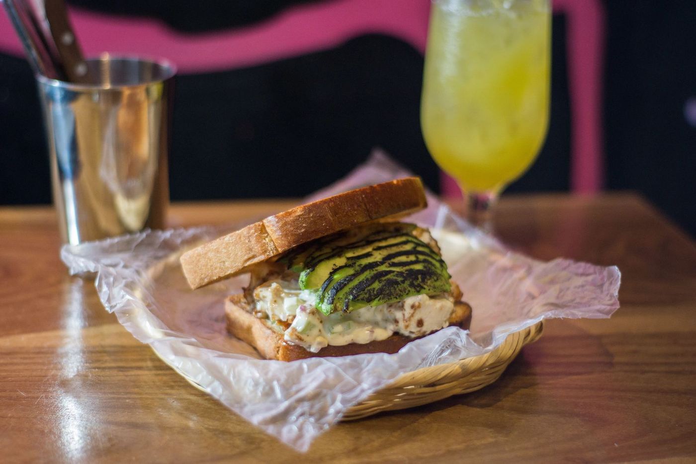 A toasted sandwich in paper on a wooden table, with a cutlery cup and a fresh drink