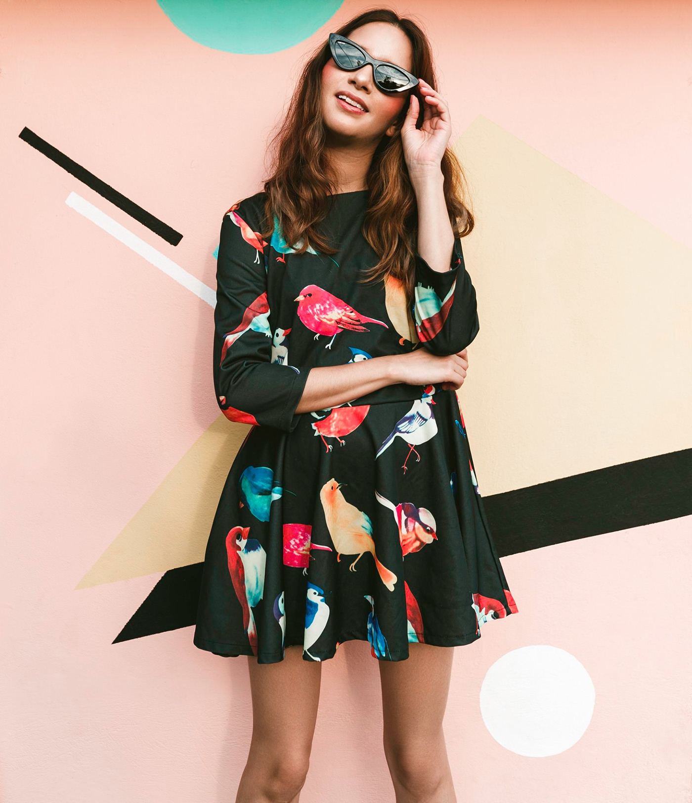 A woman in a bird-print dress and sunglasses against pastel shapes