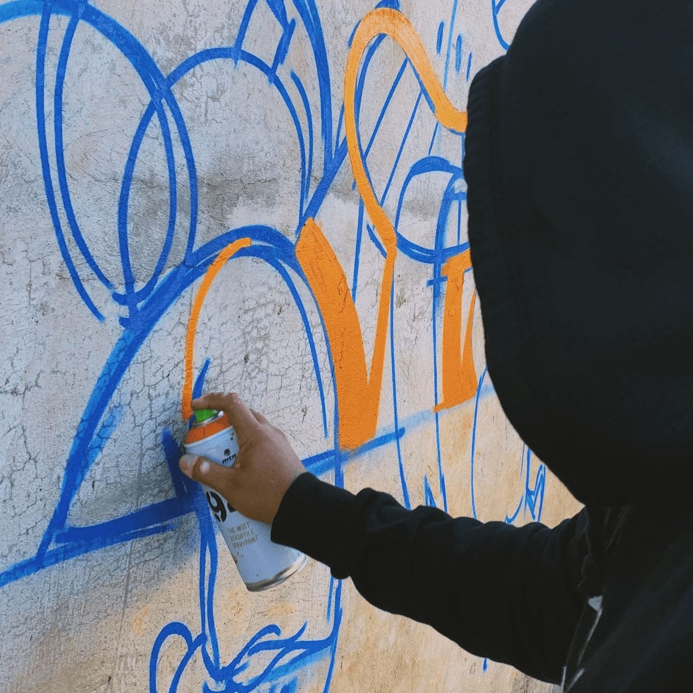 A hooded artist spraying blue and orange outlines on a wall